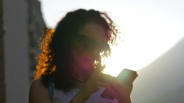 Όμορφη μελαχρινή με μακρύ φλογερό σγουρά μαλλιά σε ένα λευκό φόρεμα με τα γυαλιά είναι το περπάτημα γύρω από την πόλη και δακτυλογράφηση sms στο τηλέφωνο στο ηλιοβασίλεμα — Αρχείο Βίντεο