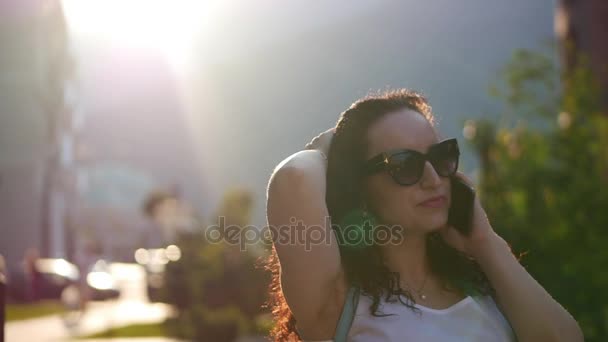 Krásná žena mluví o telefonu venku. Ona se usmívá a světlé paprsky slunce na pozadí města lze vidět v pozadí za ní. Žena s brýlemi. Zpomalený pohyb. — Stock video