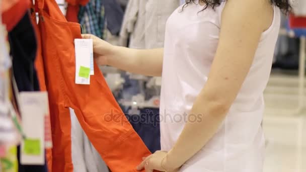 Una bella bruna sceglie un kombenizon per una piccola figlia in un negozio. Bella donna in un negozio di abbigliamento per bambini. Una bella donna compra vestiti in un centro commerciale . — Video Stock