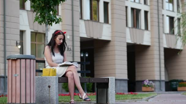 Nettes Mädchen mit rotem Rand auf dem Kopf sitzt auf einer Bank im Park und liest ein Buch. — Stockvideo