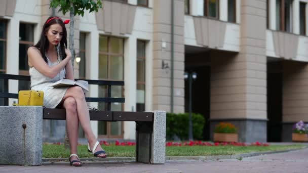 Ragazza carina con un bordo rosso sulla testa si siede su una panchina nel parco e legge un libro . — Video Stock