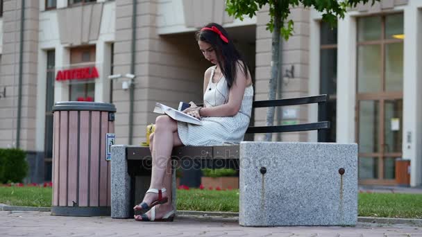 Una chica bonita con un borde rojo en la cabeza se sienta en un banco en el parque y lee el libro, luego toma el teléfono en sus manos y comienza a escribir sms . — Vídeo de stock