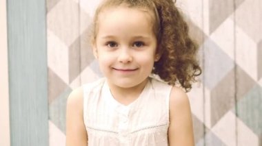 Kız bebek beyaz elbise ve kıvırcık saçlı, kameraya benziyor, tatlı tatlı gülümsedi ve biraz utangaçtır. Çocuk, çocuk, duygular.