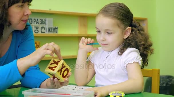 Όμορφη μικρή μαθήτρια με μια όμορφη κόμμωση και ένα όμορφο χαμόγελο για μια λευκή μπλούζα κάθεται στο γραφείο της με ένα δάσκαλο και να διδάσκει ξένες γλώσσες, να εκτελεί την εργασία ενός δασκάλου να μιλούν σωστά και να προφέρει τις λέξεις να ξέρει την προφορά και — Αρχείο Βίντεο