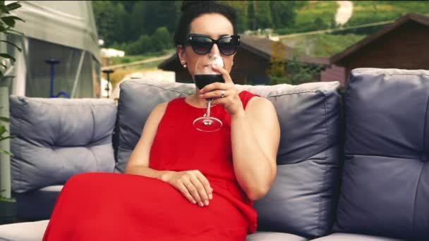 Kobieta w czarnych okularach i czerwoną sukienkę, to trzyma kieliszek czerwonego wina w dłoni, piękny widok na góry i czyste powietrze do oddychania — Wideo stockowe