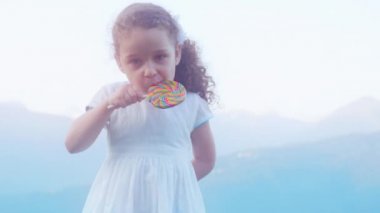 Sis çerçevede bir dağlarda beyaz elbiseli küçük güzel kız bir lolipop yiyor. 4k.