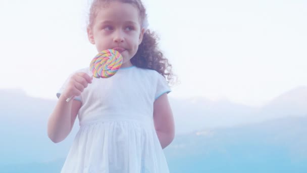 Little beautiful girl in a white dress in the mountains against a background of fog eats a lollipop. 4K. — Stock Video