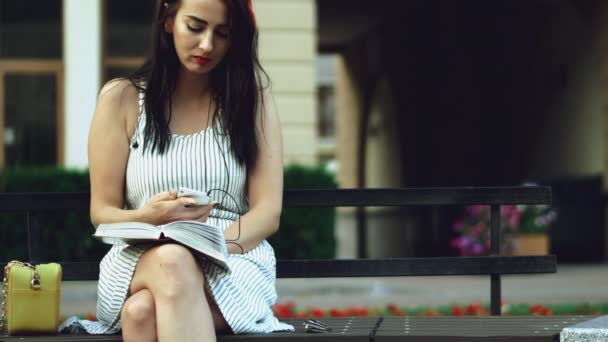 Una chica bonita con un borde rojo en la cabeza se sienta en un banco en el parque y lee el libro, luego toma el teléfono en sus manos y comienza a escribir sms . — Vídeos de Stock