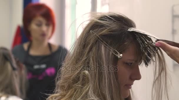 Colorazione dei capelli. Parrucchiere tintura i capelli Bionda ragazza seduta in un salone di bellezza, Barbiere spazzola capelli tiene vernice, applicare la stagnola e avvolgere i capelli, un uomo in camicia viola con un grembiule nero, specchio — Video Stock