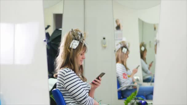 Colorazione dei capelli. Parrucchiere Vernice per i capelli è una ragazza bionda simpotico, seduto in un salone di bellezza, un parrucchiere in possesso di vernice, stampe su smythfone. 4k . — Video Stock