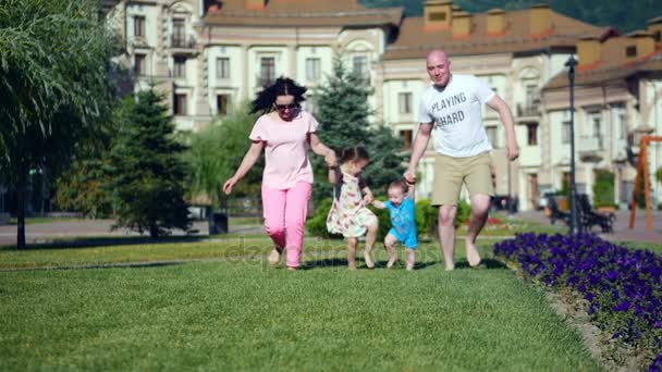 Jeune famille caucasienne insouciante court joyeusement le long de l'herbe verte tenant la main, visages heureux de la famille . — Video