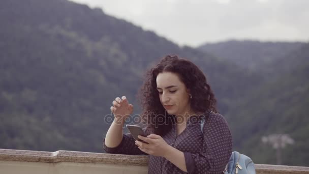 Smart, beautiful brunette corrects curly hair and prints sms on the phone at the backdrop of outstanding mountains and beautiful sunset . — стоковое видео