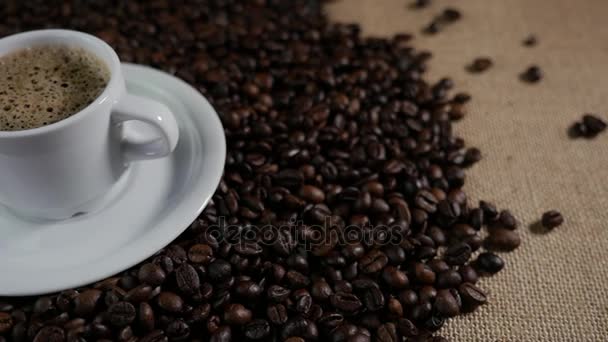 Kahve ve kahve çekirdekleri. Kahve kavrulmuş fasulye ile masada buharlaşan bir beyaz fincan. Ağır çekim kahve dökmek. — Stok video