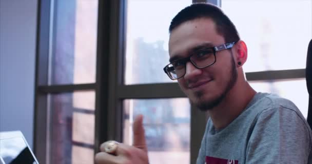 Joven feliz con gafas sentado en la mesa, haciendo trabajo en la computadora, girando y mirando a la cámara en el fondo del sol desde la ventana, sonriendo felizmente y mostrando el pulgar hacia arriba. Primer plano. 4K — Vídeo de stock