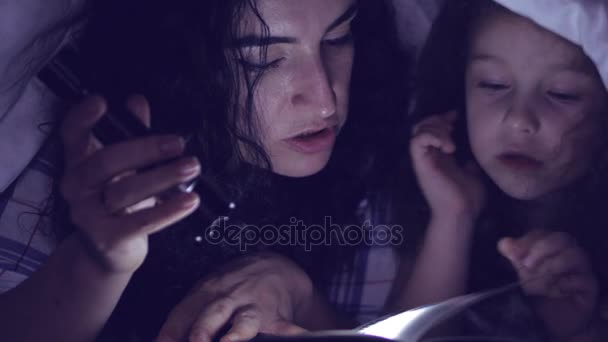 Mamá y su pequeña hija están leyendo cuentos de hadas para la noche, están envueltas en una manta caliente y sumergidas en la historia de la trama. 4K . — Vídeos de Stock