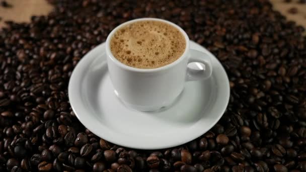 Taza de café y granos de café. Una taza blanca de espuma de café evaporada sobre una mesa con frijoles fritos . — Vídeo de stock