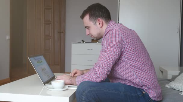 Man in werken vanuit huis met behulp van de boven- en trackpad. Jonge zakenman zittend aan tafel, koffie drinken, werk op computer. 4k — Stockvideo