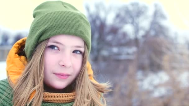 Retrato de uma jovem adolescente bonita com cabelo loiro, ao redor da neve, menina com um lenço verde e marrom em um chapéu hipster verde, sorrindo, olhando em torno de diferentes direções. Movimento lento — Vídeo de Stock