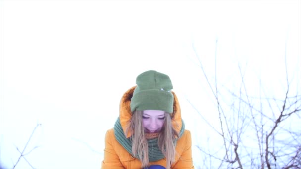 Joven hermosa adolescente adolescente con el pelo rubio, disfrutando del día de invierno al aire libre, chica con una bufanda verde y marrón en un sombrero hipster verde, arroja la nieve, mirando a la cámara. Movimiento lento — Vídeo de stock