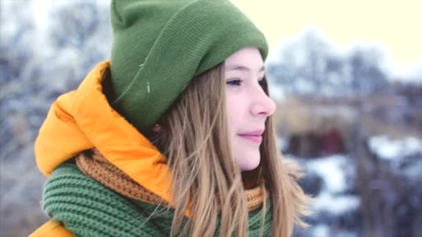 Retrato de uma jovem linda adolescente com cabelo loiro, desfrutando de dia de inverno ao ar livre, menina com um lenço verde e marrom em um chapéu hipster verde, andando ao ar livre, olhando para a câmera. Movimento lento — Vídeo de Stock