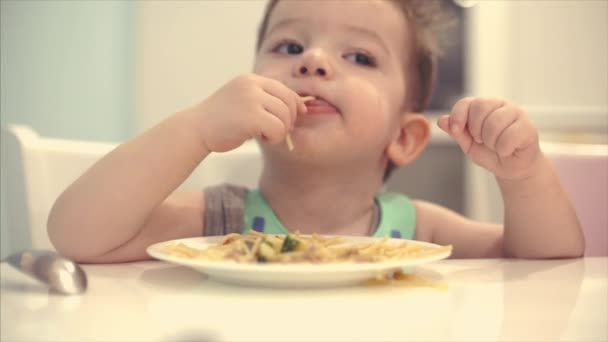 Pequeño niño está sentado en una mesa en un babero y comer sus propios espaguetis, el bebé come voluntariamente . — Vídeo de stock