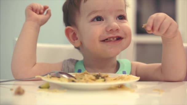 Criança pequena está sentada a uma mesa em um babador e comer seu próprio espaguete, o bebê come de bom grado. Instrução . — Vídeo de Stock