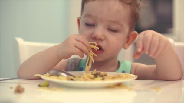 Kleines Kind sitzt im Lätzchen am Tisch und isst seine eigenen Spaghetti, das Baby isst bereitwillig. — Stockvideo