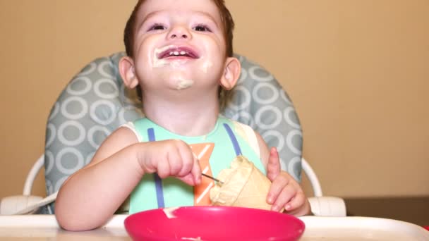 Piccolo bambino si siede dietro un seggiolone per, in un pettorale e c'è un gelato in un bicchiere, il bambino sorride alla bocca piena. Rallentatore . — Video Stock