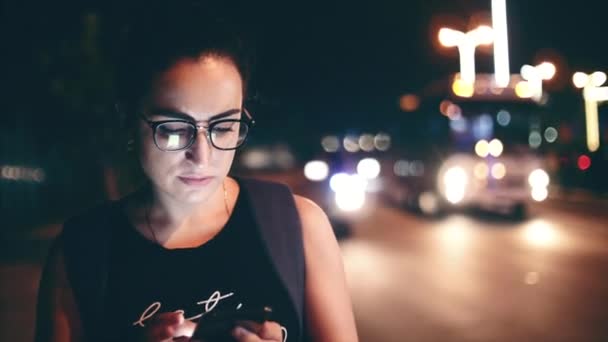 Wanita yang menarik menggunakan ponsel saat berjalan di jalan-jalan di kota malam hari, di latar belakang dapat melihat pengendara motor. Rekaman stok . — Stok Video