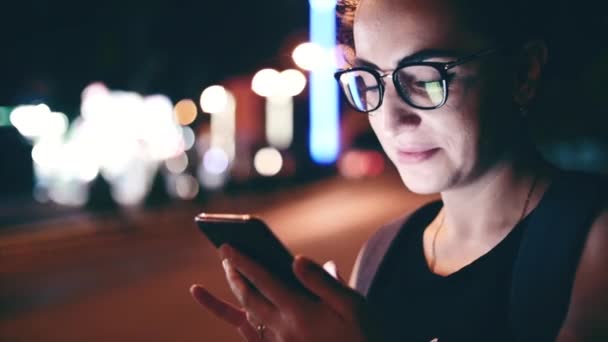 Mujer atractiva usando un teléfono móvil mientras camina por las calles en una ciudad nocturna, en el fondo se puede ver a los ciclistas. Material de archivo . — Vídeos de Stock