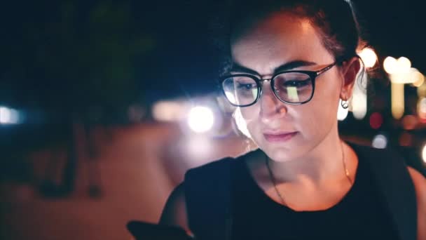 Mujer atractiva usando un teléfono móvil mientras camina por las calles en una ciudad nocturna, en el fondo se puede ver a los ciclistas. Material de archivo . — Vídeos de Stock