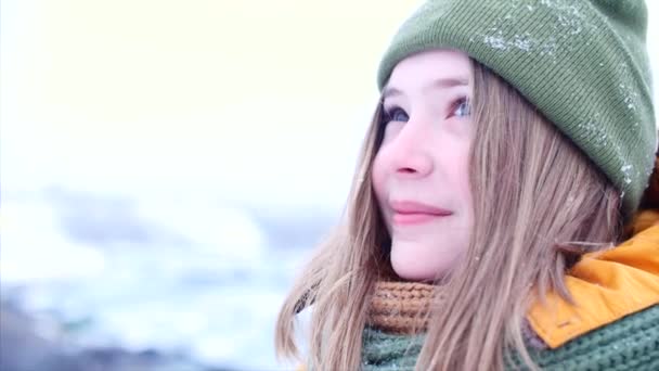 Young beautiful teenage girl teenage with blond hair, enjoying winter day outdoors, girl with a green and brown scarf in a green hipster hat, throws up the snow, looking at the camera. Slow motion — Stock Video
