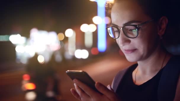 Attractive woman using a mobile phone while walking through the streets in a night city, in the background can see bikers. Stock footage. — Stockvideo