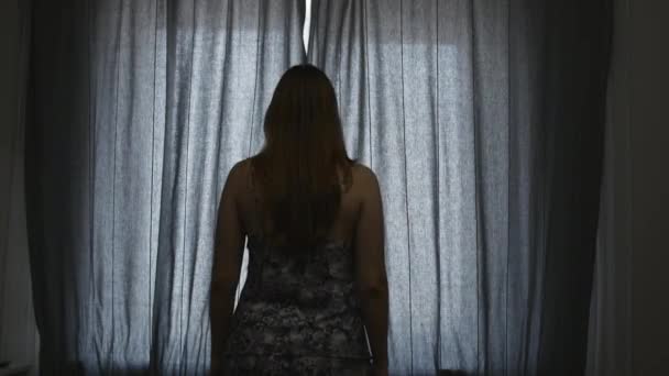 Jonge vrouw onthullen gordijn en kijken uit het venster. — Stockvideo