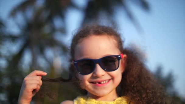 Happy babymeisje in een gele jurk en zonnebril, krullend haar, kijkt naar de camera, lachend en een beetje verlegen. Kind, kinderen, emoties. — Stockvideo