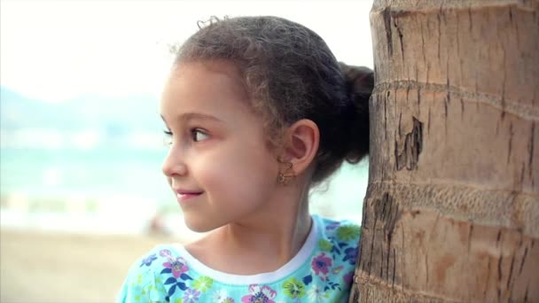 Happy little girl on the beach in a blue dress with a palm tree, looking at the camera, smiling and a little shy. Child, children, emotions. — Stock Video