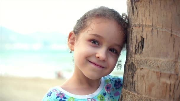 Menina feliz na praia em um vestido azul com uma palmeira, olhando para a câmera, sorrindo e um pouco tímido. Criança, crianças, emoções . — Vídeo de Stock