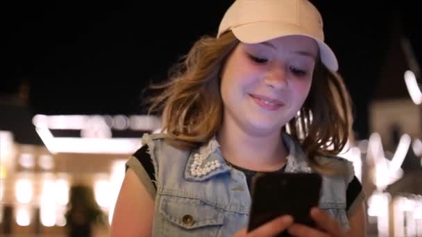 Aantrekkelijke tienermeisje met behulp van een mobiele telefoon terwijl het lopen door de straten in een stad van de nacht, in de stad van de nacht achtergrond. Beeldmateriaal. — Stockvideo