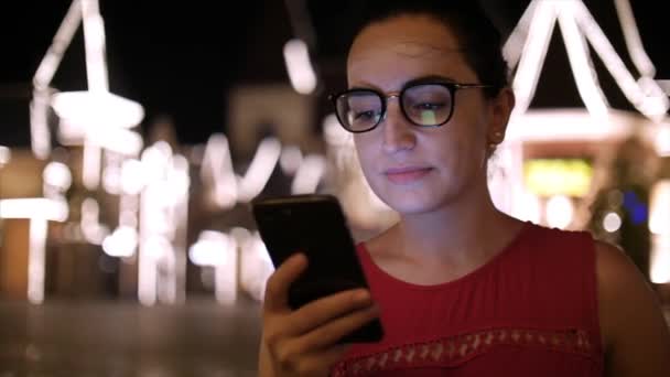 A vonzó lány a mobiltelefon használata egy éjszaka városban, a háttér éjszakai város utcáin sétálva. Stock footage. — Stock videók