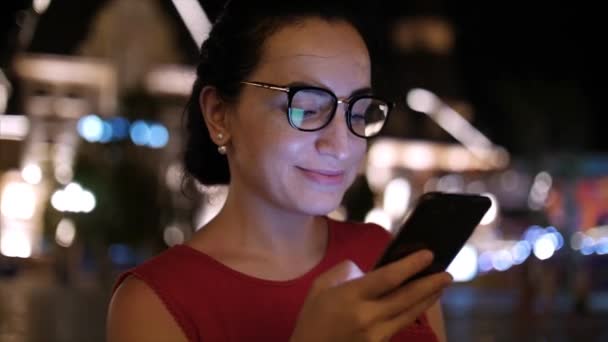Femme attrayante utilisant un téléphone portable tout en marchant dans les rues dans une ville de nuit, dans la ville de nuit de fond. Images de stock . — Video