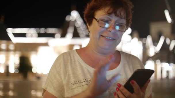 Vonzó nő használ egy mozgatható telefon egy éjszaka városban, a háttér éjszakai város utcáin sétálva. Stock footage. — Stock videók