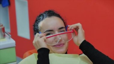 Kadın diş hekimi klinikte kadın hastayı tedavi ediyor. Dişçi resepsiyonundaki hasta güvenlik gözlüğü takıyor, kameraya bakıyor ve gülümsüyor. Diş muayenesi kavramı.