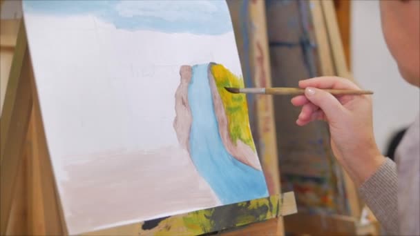 Τα χέρια του καλλιτέχνη, μια γυναίκα καλλιτέχνης ζωγραφίζει έναν καμβά με ένα πινέλο, κάθεται σε ένα καβαλέτο και κάνει πινελιές στον καμβά. Σχέδιο διαδικασία: στο εργαστήριο τέχνης. Σκίτσο σε καμβά. — Αρχείο Βίντεο