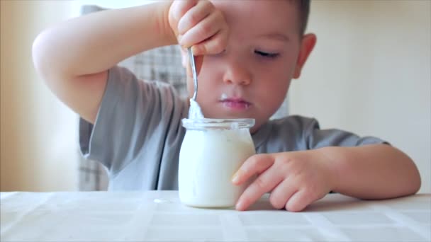 Zbliżenie dziecka siedzącego przy stole i jedzącego jedzenie dla niemowląt łyżeczką. 2-3-letni chłopiec chętnie je jogurt mleczny lub kefir, śmietanę, jedzenie dla dzieci. 4 K — Wideo stockowe