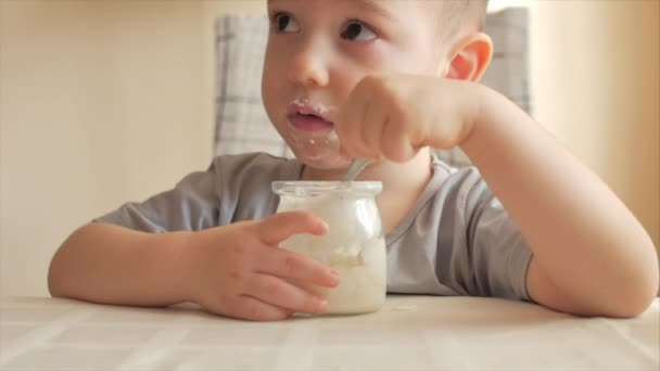 Close-up de um bebê sentado à mesa e comendo comida de bebê com uma colher por conta própria. Um menino de 2-3 anos come ansiosamente iogurte de leite ou kefir, creme azedo, comida para bebês. 4 K — Vídeo de Stock