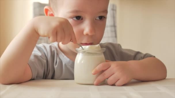 Mały chłopiec siedzący przy stole i jedzący jedzenie dla niemowląt łyżeczką. 2-3-letni chłopiec chętnie je jogurt mleczny lub kefir, śmietanę, jedzenie dla dzieci. — Wideo stockowe