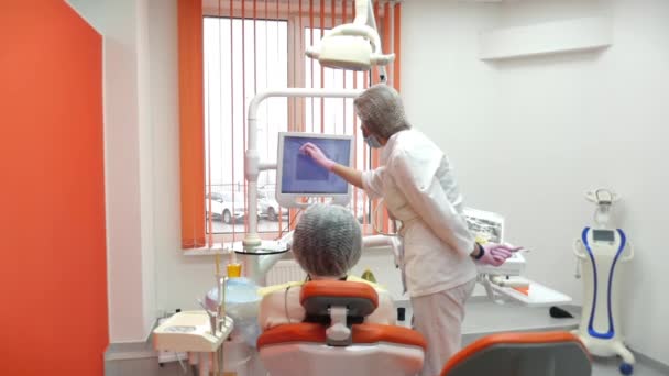 Tandarts trakteert vrouwen op een patiënt in de kliniek. Tandarts toont de patiënt op de tablet hoe de tanden zullen worden behandeld, geeft lessen over de juiste verzorging van de tanden en de mondholte. — Stockvideo
