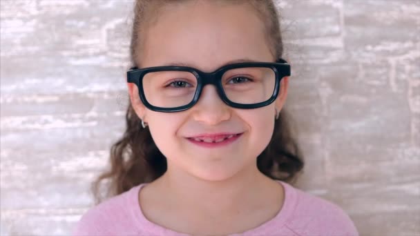 Portret van een leuk meisje, gelukkig kind dat glimlachend naar de camera kijkt. Klein meisje met een zwarte bril en een roze trui. — Stockvideo