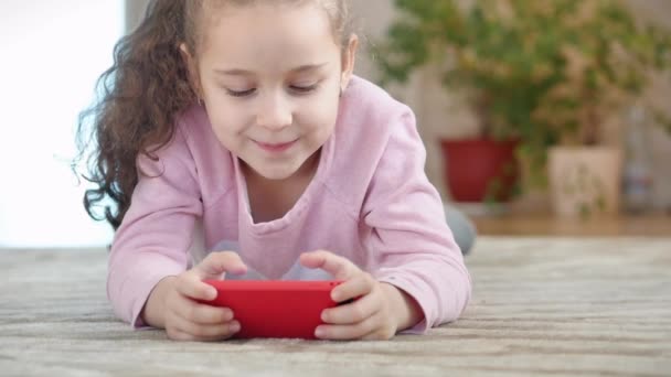 携帯電話やタブレットで楽しいかわいい赤ちゃん。小さな女の子は、モバイルゲームをプレイする彼女の自由な時間を費やして、彼女の手で明るい画面をクラッシュさせます。コンセプト:子供のための幸せな子供時代、ゲーム. — ストック動画