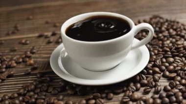 Kahve fincanı ve kahve çekirdeği. Tahta bir masanın üzerinde beyaz bir fincan kahve ve kahve çekirdekleri duruyor, yakın plan kahve damlaları bir fincanın içine düşüyor..
